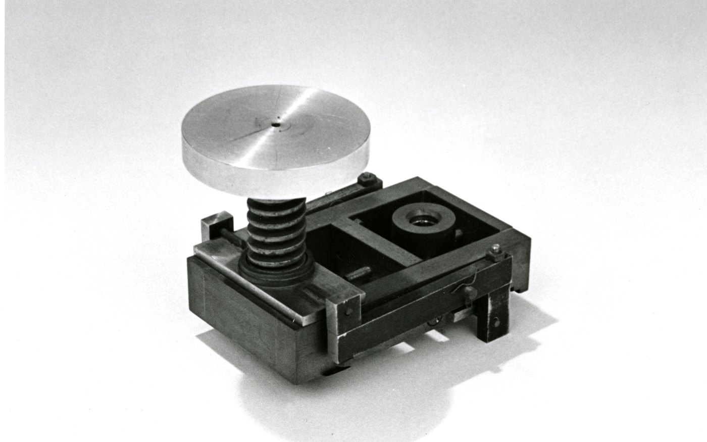 An early diamond anvil cell, a small steel frame with a spring-loaded screw press and a polished metal disc on top, photographed against a plain background