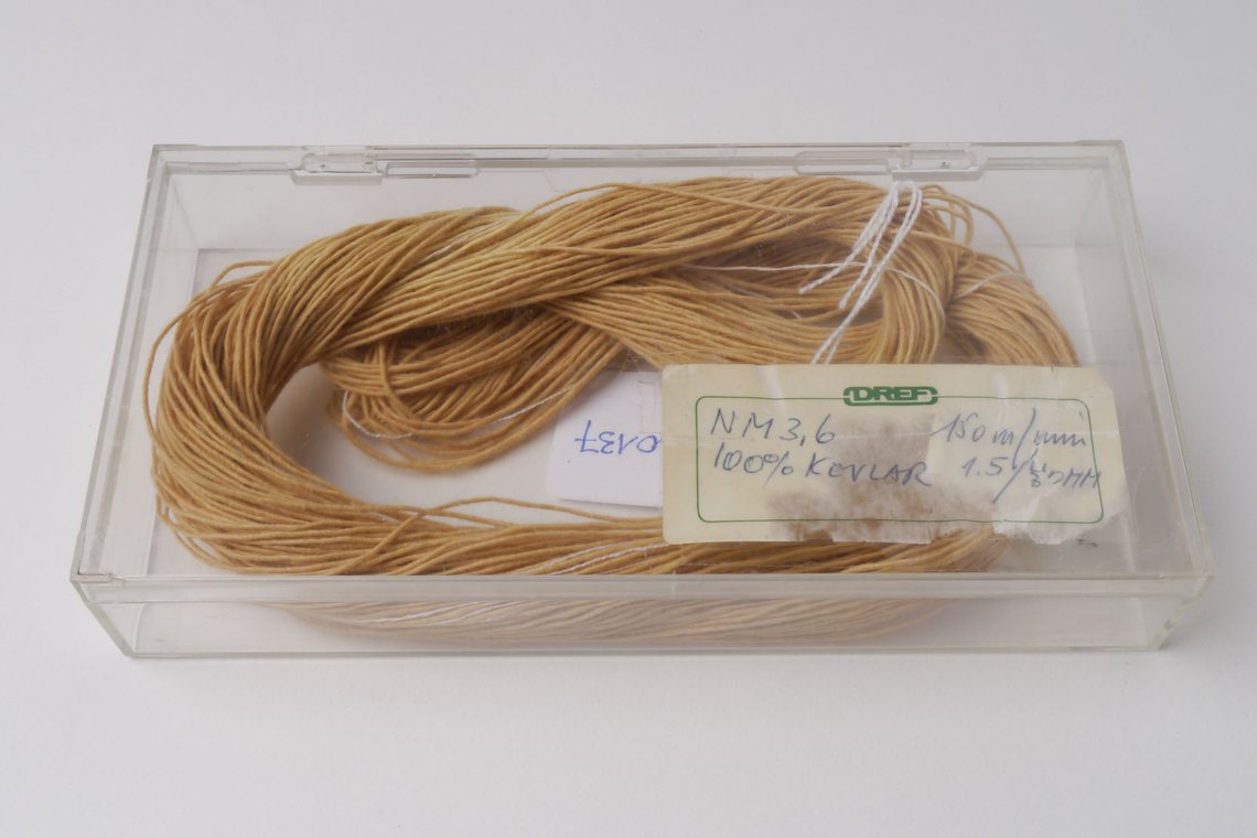 Real sample of Kevlar yarn: a hank of fine golden-tan aramid filament coiled inside a clear plastic museum specimen box, with a handwritten label reading “NM 3,6 — 100% KEVLAR”.