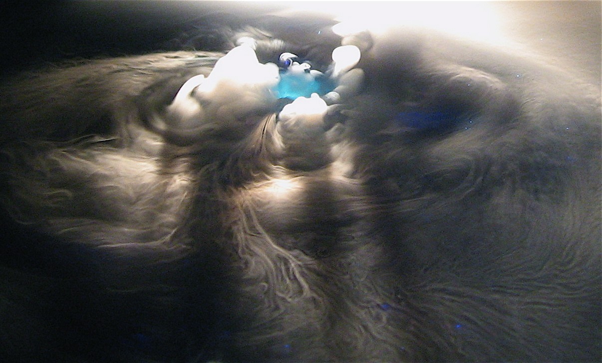 Dry ice subliming on a water surface, the fog folding into spirals and a pale blue glow at the centre